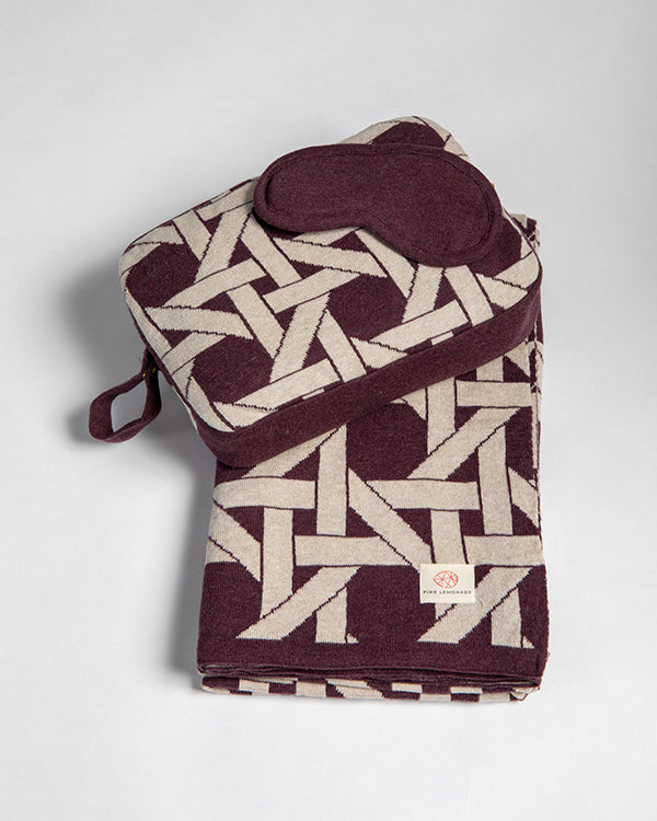 Folded burgundy blanket with geometric pattern and matching eye mask on a light gray background.