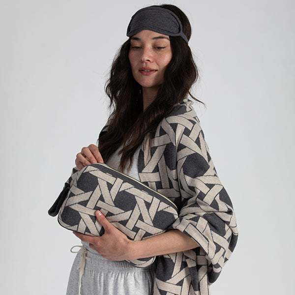 Woman holding a grey patterned pouch and wearing a matching blanket and eye mask on a plain background.