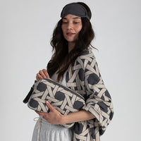 Woman holding a grey patterned pouch and wearing a matching blanket and eye mask on a plain background.