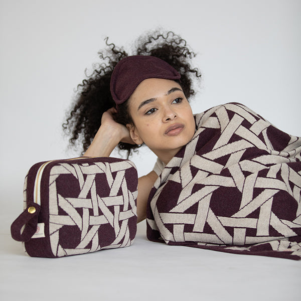 Woman wearing a burgundy eye mask and holding a matching pouch and blanket with a geometric design on a plain background.