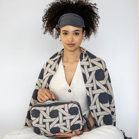 Woman wrapped in a geometric patterned grey blanket holding a matching pouch with a plain background.