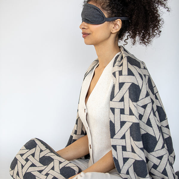 Woman wearing a patterned blanket and grey eye mask on a plain background.