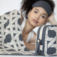 Woman lying on couch with a patterned grey blanket and eye mask with a matching pouch next to her.