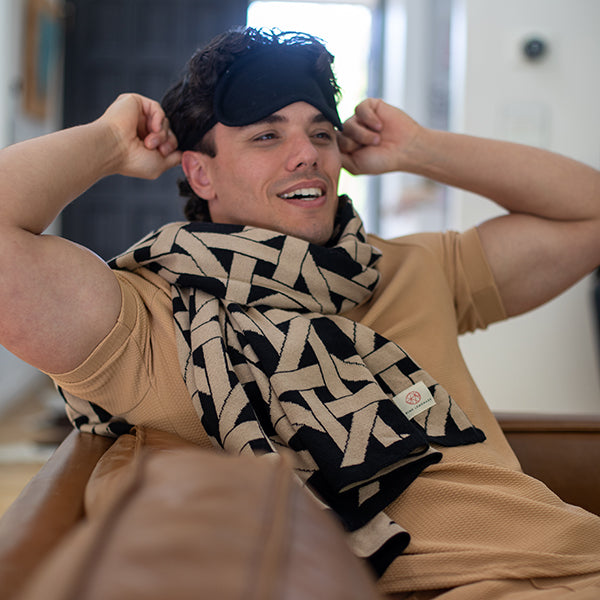 Man wearing a black and beige patterned geometric blanket around his neck and a sleep mask, sitting back on a couch.