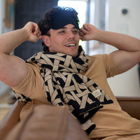 Man wearing a black and beige patterned geometric blanket around his neck and a sleep mask, sitting back on a couch.