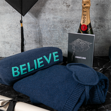 Cotton navy blue blanket with 'BELIEVE' text, bottle of Moët champagne, and graduation card on a dark surface.