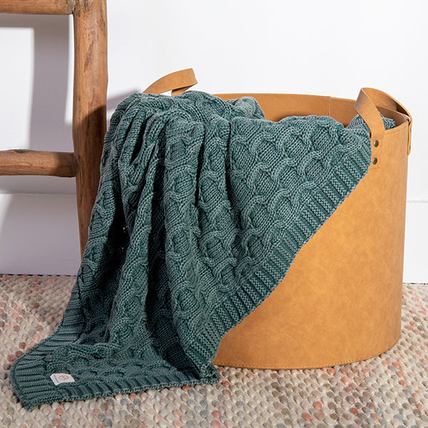 Green knitted textured cotton blanket draped over a brown basket on a textured mat.