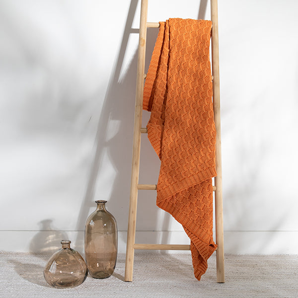 Orange textured cotton blanket draped over a wooden ladder against a white wall with two glass bottles on the floor.