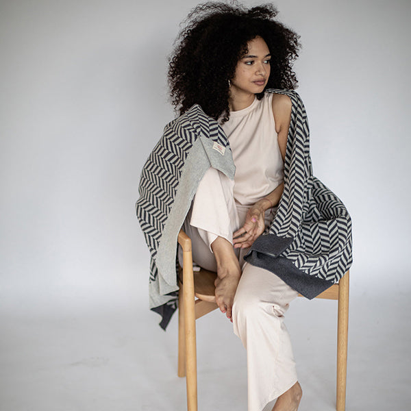 Woman sitting on a wooden chair wearing a dark gray herringbone patterned throw against a plain background.