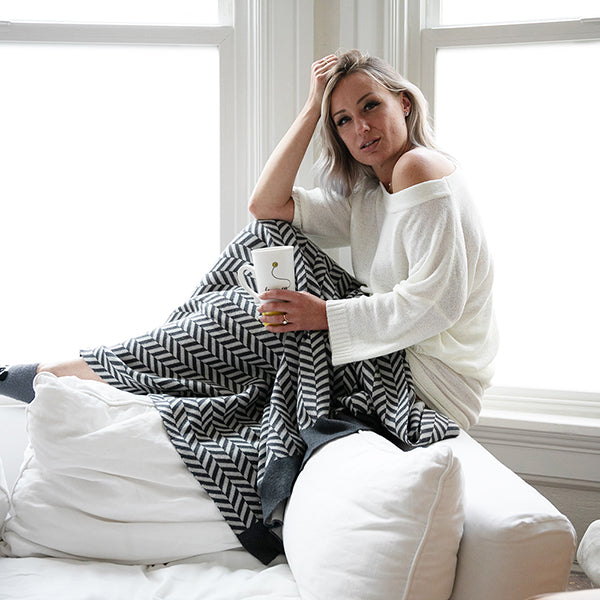 Woman sitting on a white couch with a grey cotton blanket with a herringbone design and a mug, in a bright room with large windows.