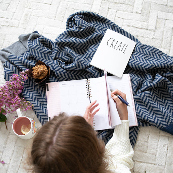 Child sitting on a white rug with a navy blue herringbone patterned blanket, writing in a notebook with a cup of lemonade.